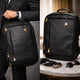 Black leather laptop bag held by a person in a suit, with another backpack on a marble surface.